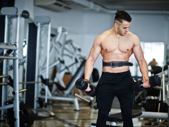 Muscular arab man training in with dumbbells modern gym. Fitness arabian men with naked torso doing workout .